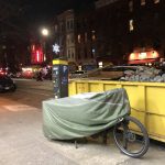 Covered cargo bike (olive cover) next to a yellow dumpster