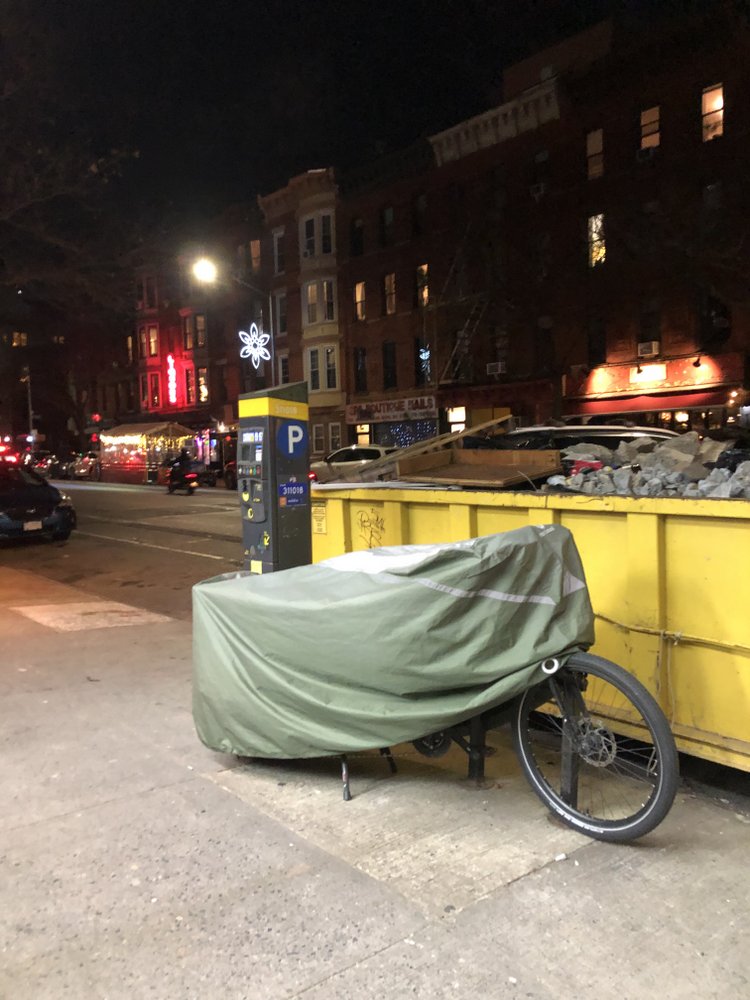 Covered cargo bike (olive cover) next to a yellow dumpster