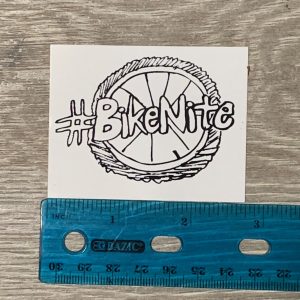 #BikeNite sticker (white) on grey background with ruler for scale. 2.75" wide