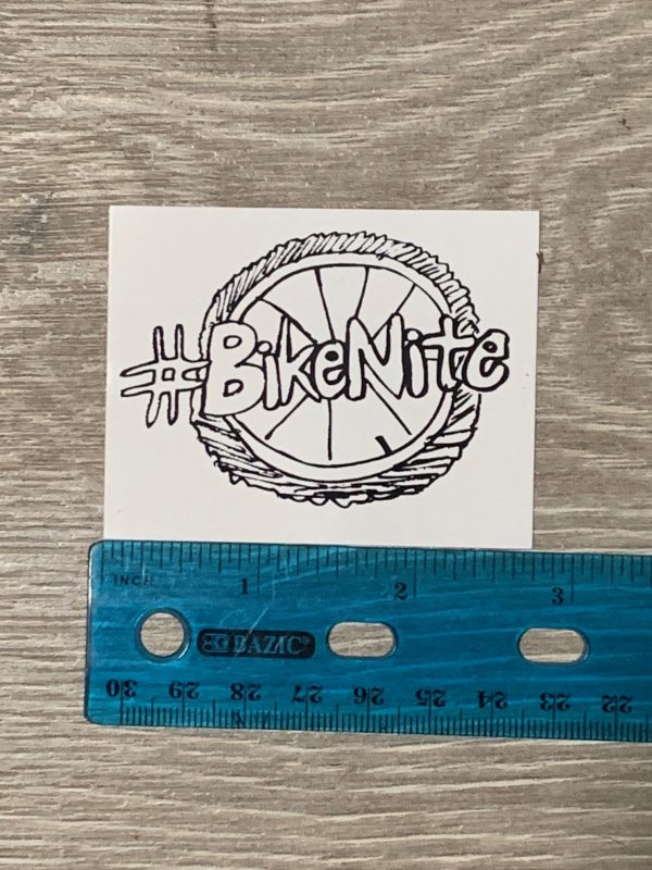 #BikeNite sticker (white) on grey background with ruler for scale. 2.75" wide